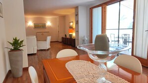 Un comedor con una mesa lista para dos personas, vista despejada de la sala de estar al fondo y un amplio ventanal con cortinas.