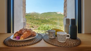 Ontbijt met croissants, een banaan en twee mokken op een tafel bij een raam met uitzicht op een bergachtig landschap.