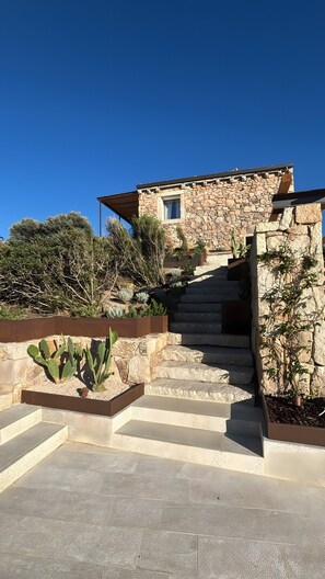 Une maison en pierre avec un escalier menant à l’entrée, entourée d’une végétation désertique.