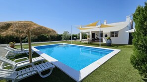 En pool med vita solstolar, ett parasoll med halmtak och ett modernt hus med vit fasad.