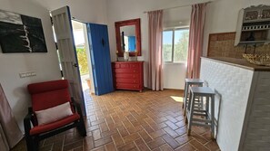 Ett rum med en röd stol, en blå dörr, en röd byrå, en vit kakelbeklädd köksbänk och ett fönster med gardiner.