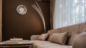 A cozy living room with a brown sofa, a round wall clock, and a modern light fixture. - Second home Next to Sea Garden (Varna)