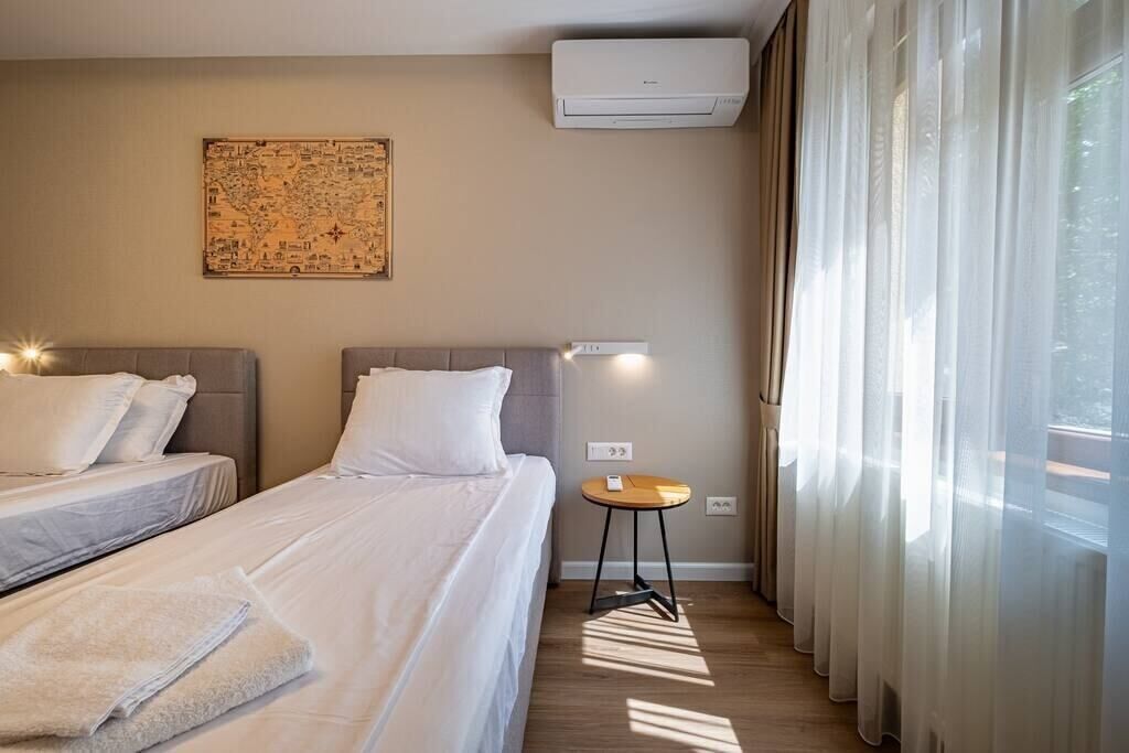 A hotel room with two beds, a wall-mounted air conditioner, a small table with a lamp, and sheer curtains allowing natural light.