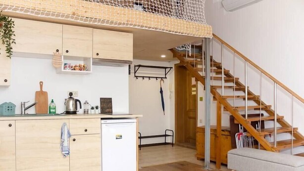 Une cuisine compacte avec des meubles en bois, un petit réfrigérateur et un étendoir suspendu. Un escalier mène à un espace en mezzanine doté d’un plafond en treillis.