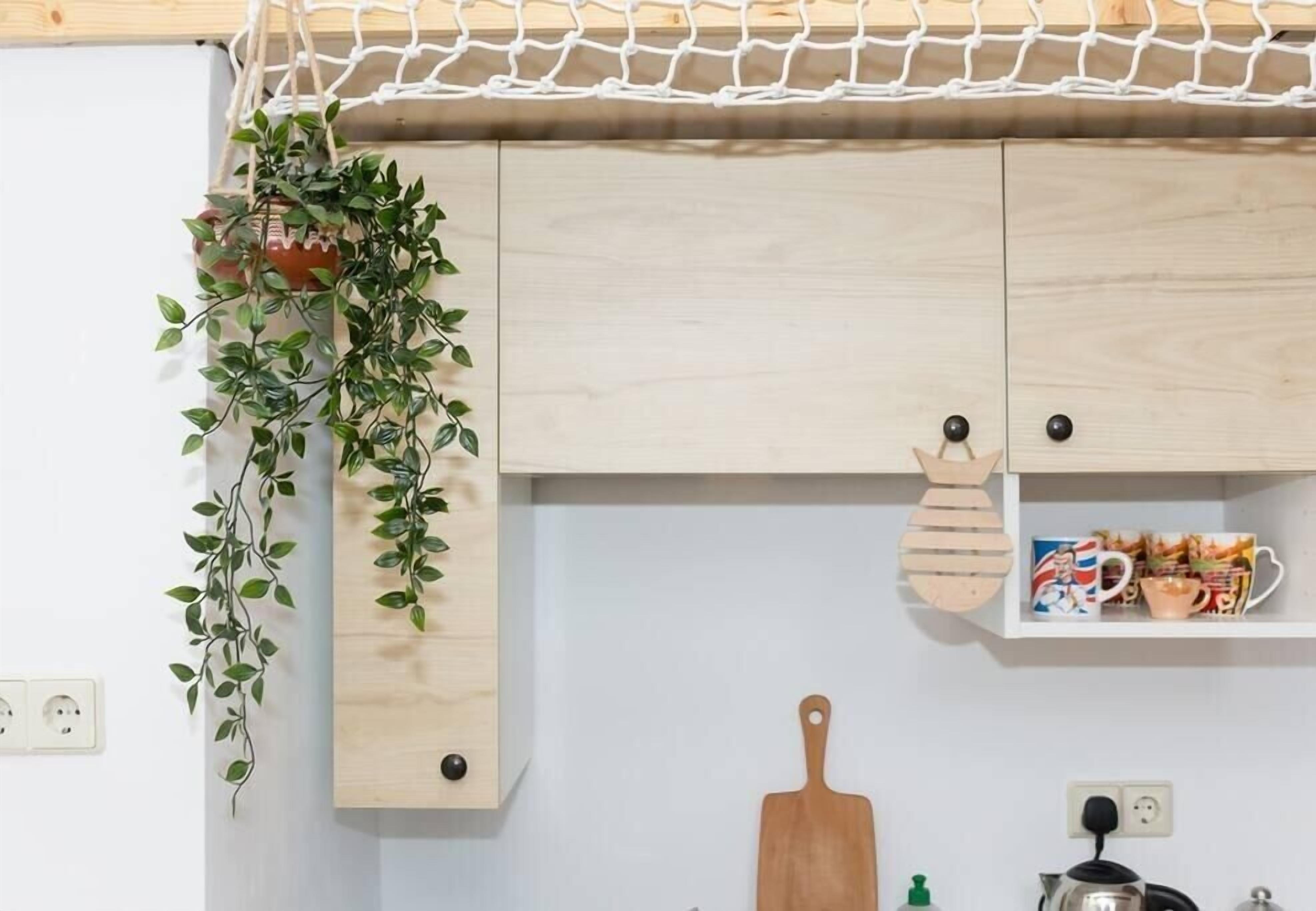 Une cuisine avec des meubles de rangement en bois, une plante suspendue, une planche à découper en bois et une étagère où sont posées des tasses.