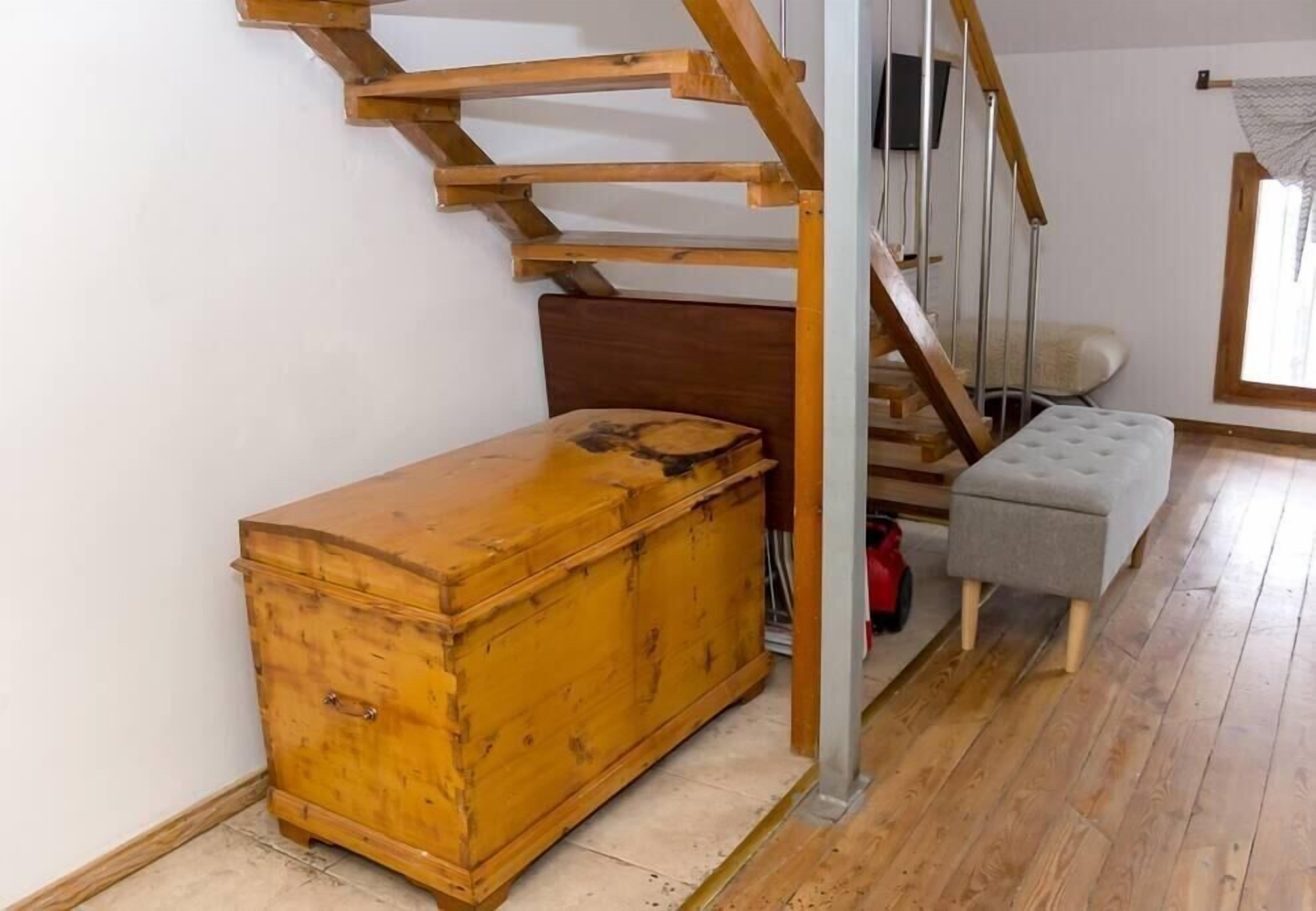 A wooden chest of drawers, a grey ottoman, and a staircase with wooden railings in a room with wooden flooring.