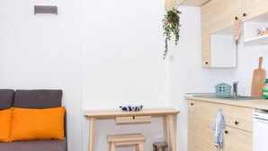 Un espace de vie compact comprenant un canapé gris, un bureau en bois avec un tabouret, une petite poubelle, un coin cuisine avec un évier et une plante suspendue.