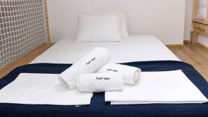 Un lit bien fait, avec une literie et des oreillers blancs, une couvre-lit bleue et trois serviettes blanches roulées, toutes portant la marque « FLAT WAY ».