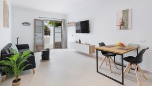 Una sala de estar moderna con un sofá, una mesa de comedor con sillas, un televisor montado en la pared y un ventanal corredero que da a un espacio exterior.