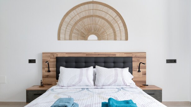 Una cama bien tendida con una colcha estampada, dos mesitas de noche con lámparas y un adorno en la pared sobre la cama.