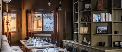A dining area with a long table set for six, a bookshelf, and a view of a snowy landscape.