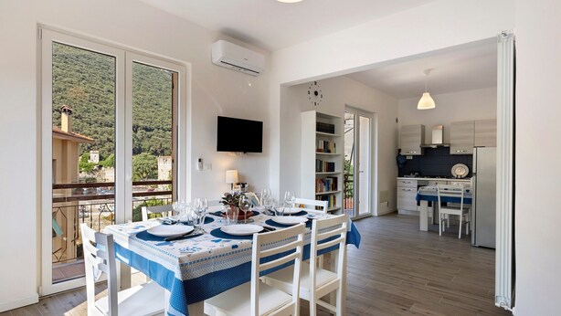 A dining area with a table set for six, a kitchen in the background, and a balcony with a view of a hillside. - Apartment 'Il Borgo Incantato with Garage' with Wi-Fi and Air Conditioning (Finale Ligure)