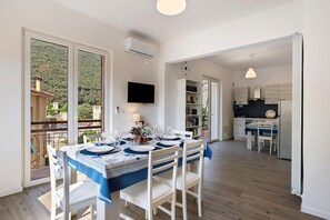 A dining area with a table set for six, a kitchen in the background, and a balcony with a view of a hillside.
