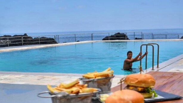 Un espace repas aménagé au bord de la piscine, offrant une vue sur l’océan, avec des hamburgers et des frites servis à table.