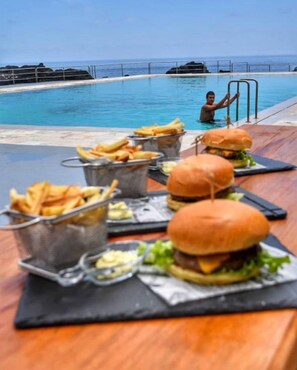 Spiseområde ved poolen med udsigt over havet, hvor der serveres burgere og pommes frites.