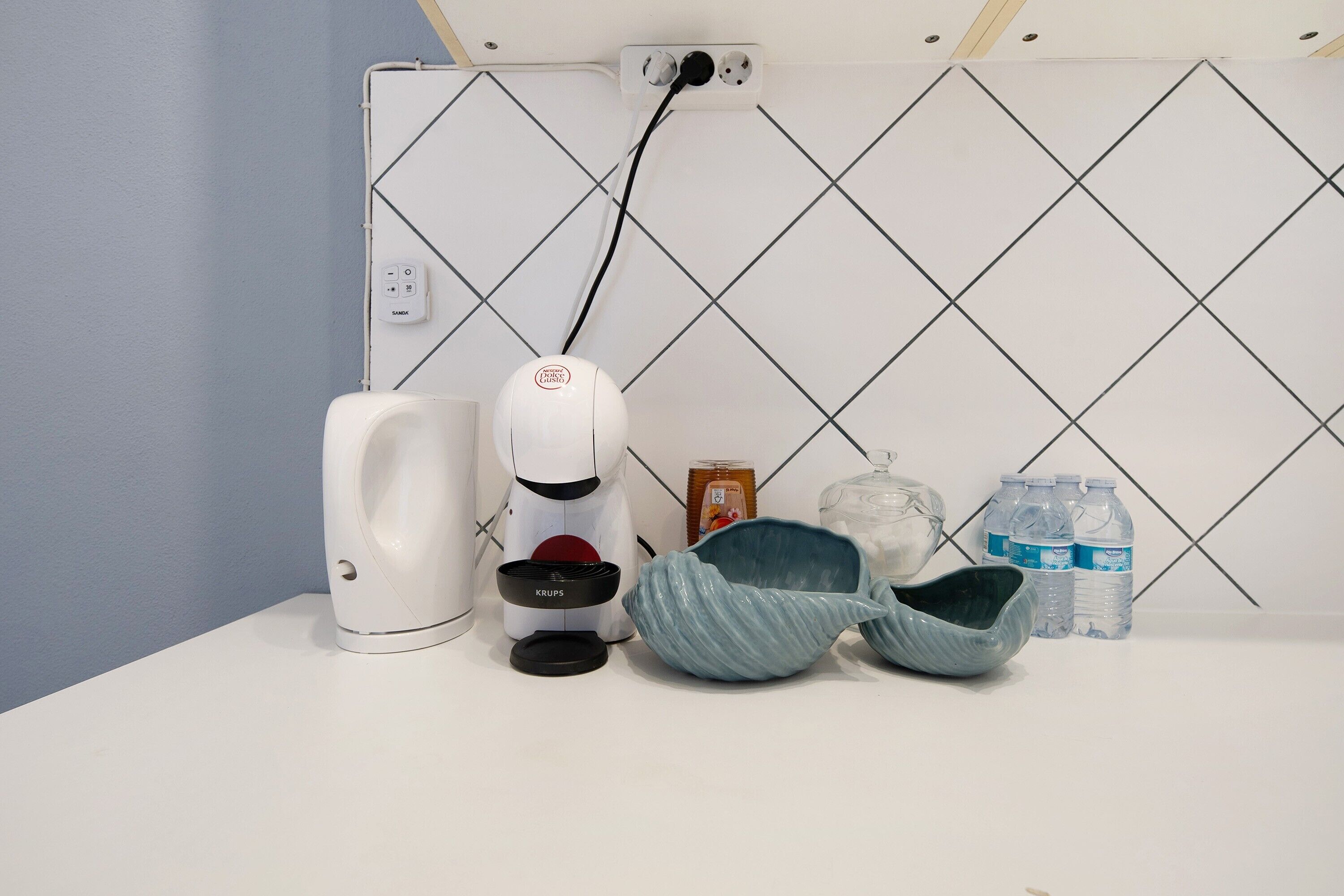 Et badeværelse med et hvidt toilet, et hvidt bidet, en Krups eltandbørste, en blå keramikvask og vandflasker på bordet.