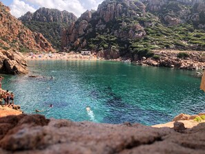 A coastal beach with clear water, rocky terrain, and a mountainous backdrop.
