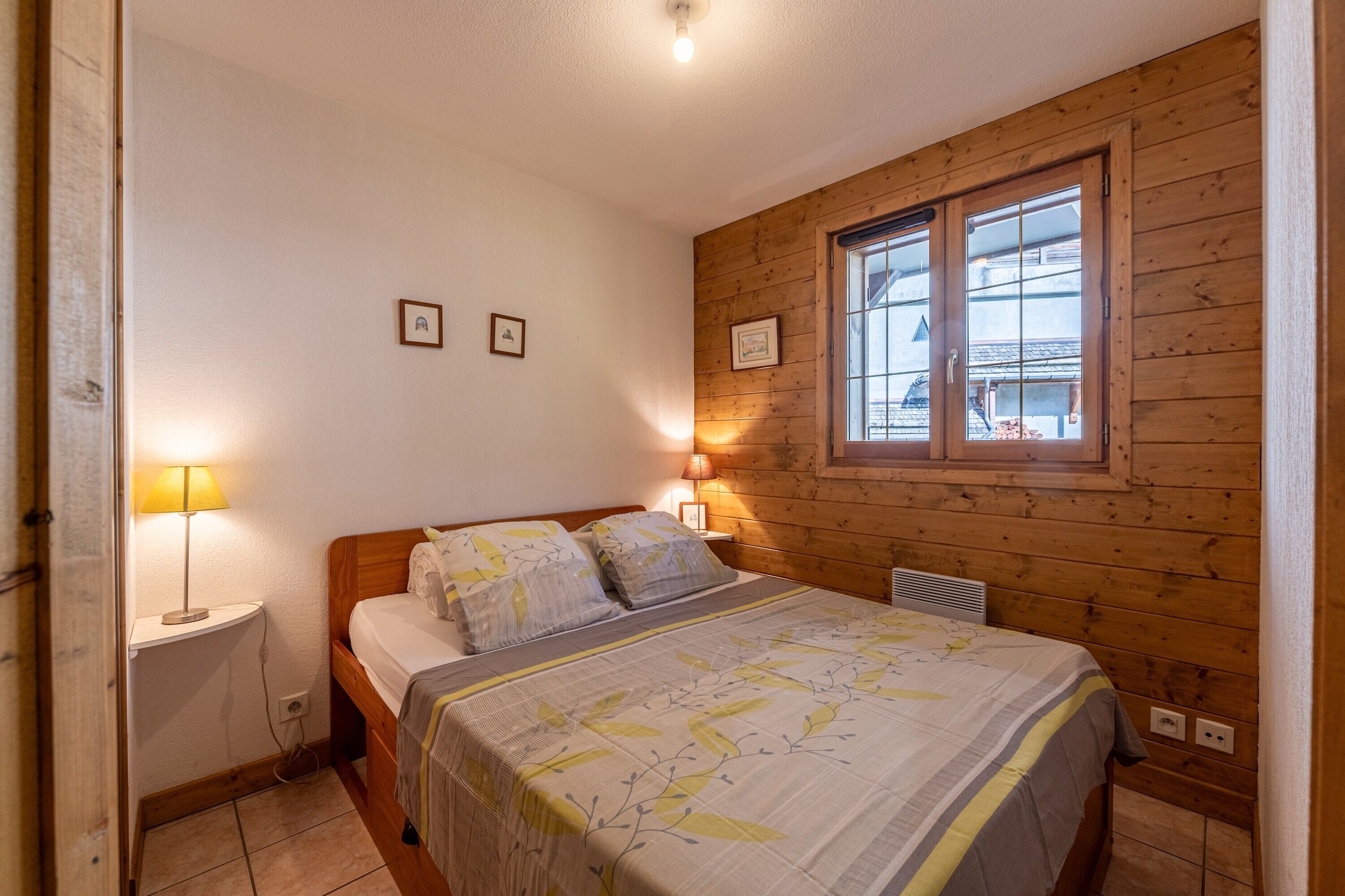 A bedroom with a wooden wall, a bed with a floral bedspread, two bedside lamps, and a window with a view.