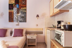 A compact hotel room with a bed, bedside table, a small kitchen with a stove and microwave, and a wall-mounted abstract painting.