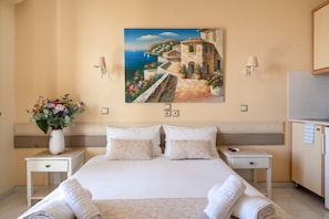 Quarto de hotel com cama, duas mesinhas de cabeceira, um vaso de flores, um quadro com uma paisagem costeira e vista para o mar.
