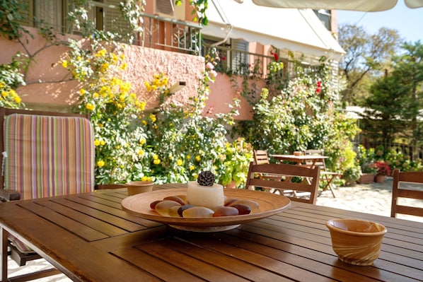 Une table en bois, sur laquelle se trouvent une assiette de nourriture et un bol, entourée de chaises et d’un bâtiment orné de plantes.