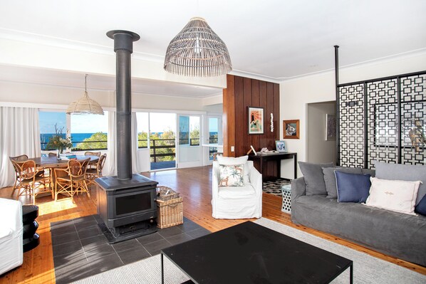 A spacious living room with a large black stove, a grey sofa, and a dining area with wicker chairs. - Bannisters Beach House (Mollymook Beach)