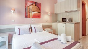 A hotel room with a bed, a small kitchenette, a painting on the wall, and two wall-mounted lamps.