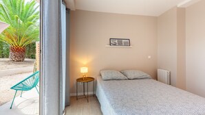 A bedroom with a bed, a bedside table with a lamp, a framed picture on the wall, and a view of a palm tree outside. - Apartment 'Havre De Paix' with Shared Pool, Private Terrace and Wi-Fi (Marseille)
