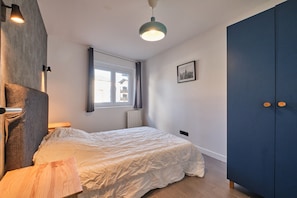 Un dormitorio con cama, ventana con cortinas, lámpara de pared, radiador y armario.