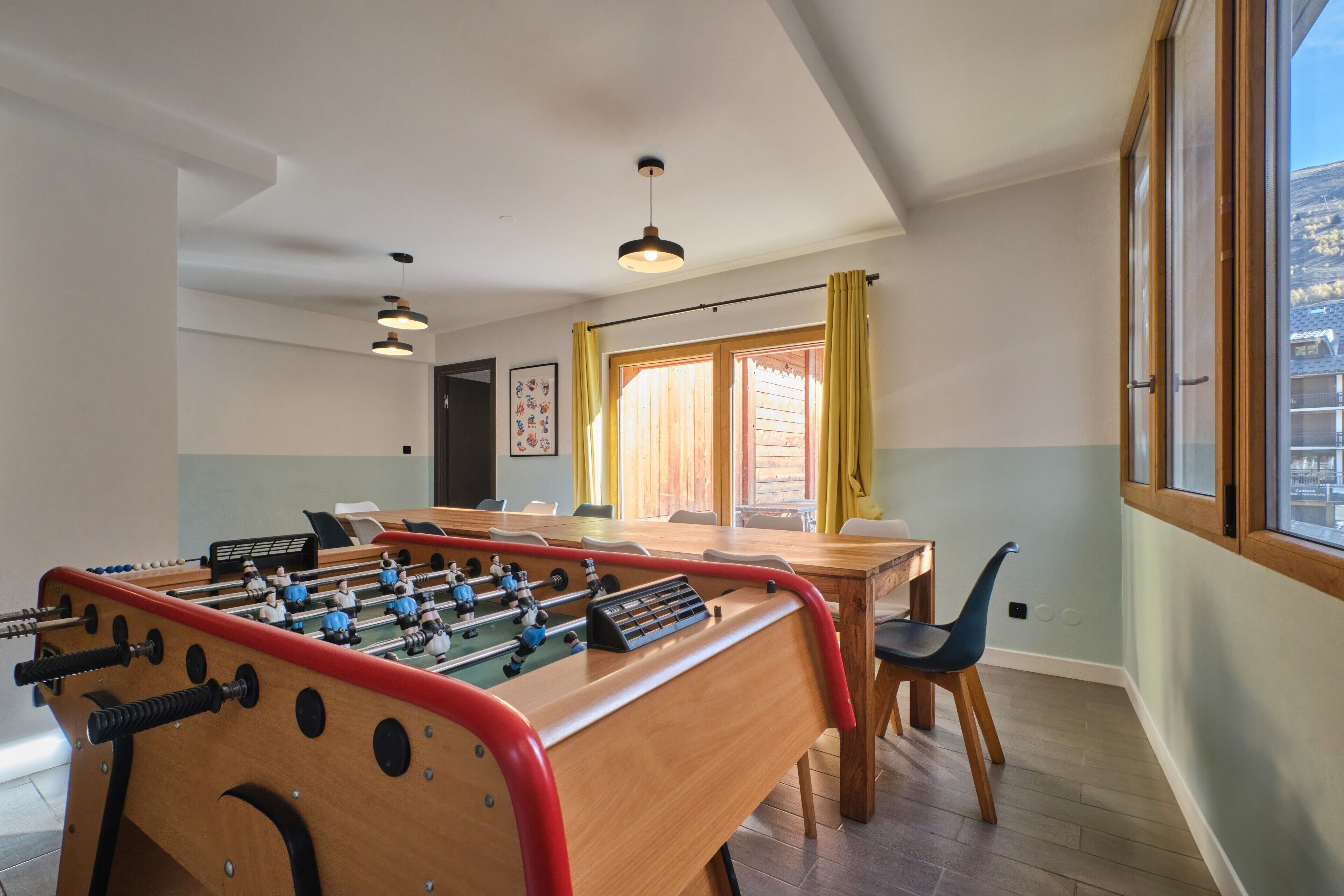 A room with a foosball table, a dining table, chairs, a window with curtains, and a ceiling light.