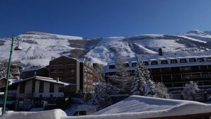 Una montagna innevata con una seggiovia, edifici con balconi e un cielo azzurro limpido.