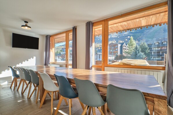 Un espace repas moderne avec une longue table en bois et des chaises, de grandes fenêtres avec des rideaux et un téléviseur à écran plat fixé au mur.