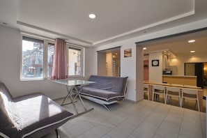 Ein modernes Wohnzimmer mit einer Couch, einem Glastisch, einem Essbereich mit Stühlen und einer Küche im Hintergrund.