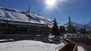 Paisaje montañoso nevado con un sol brillante, un edificio con varias ventanas y una valla de madera.