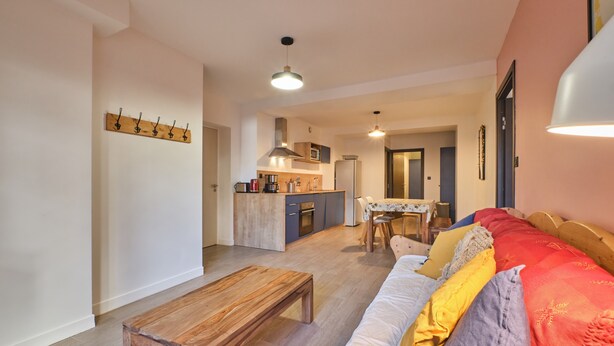 A living room with a wooden coffee table, a sofa with colorful cushions, a kitchen area with a dining table, and a coat rack.