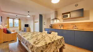A bedroom with a wooden bed, a dining table with a floral tablecloth, a kitchen with a microwave, and a window with yellow curtains.