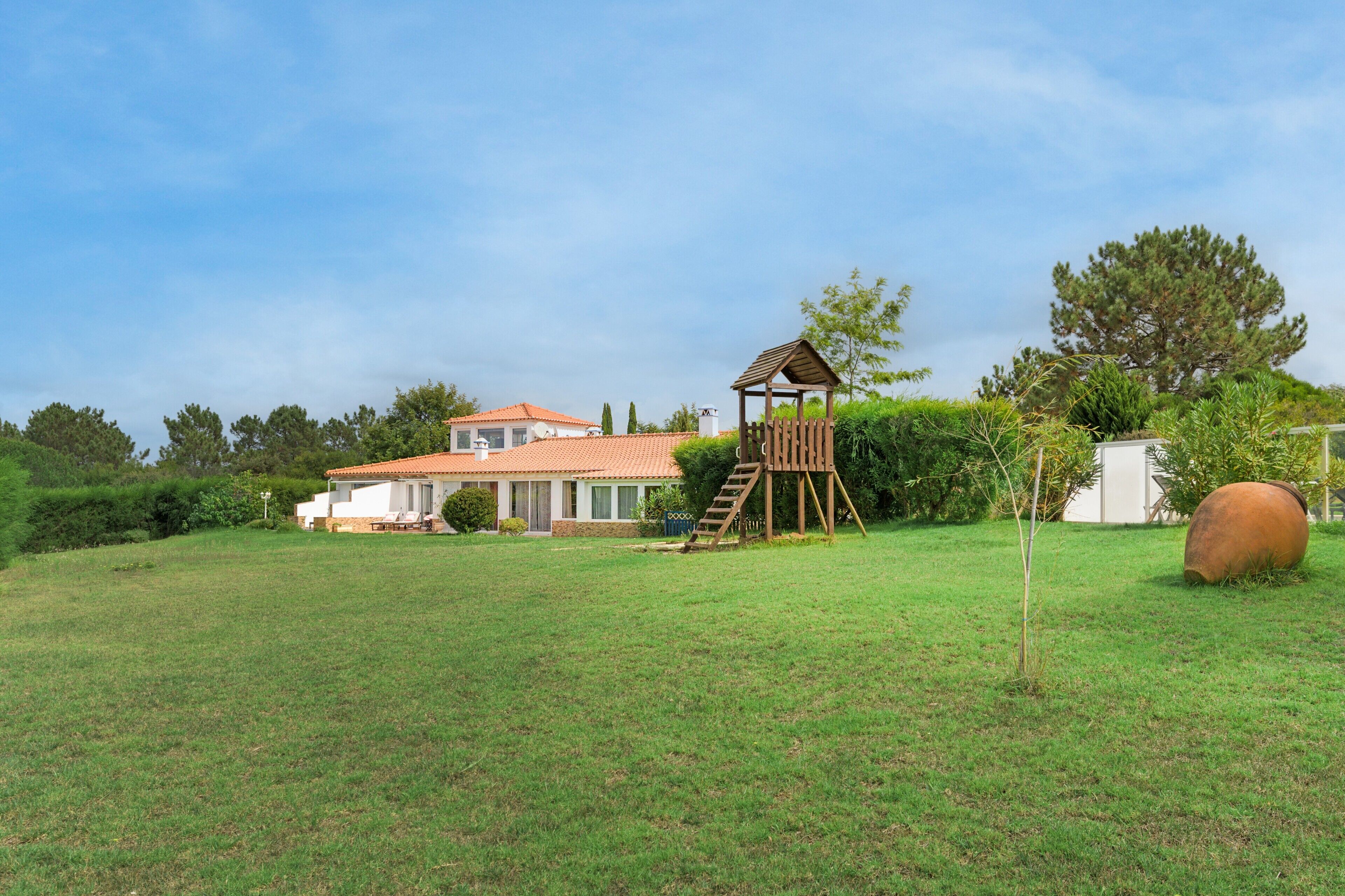 Una casa con techo de tejas rojas, un parque infantil de madera y un objeto esférico grande en el jardín.