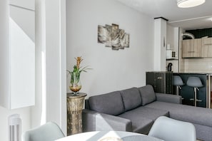 Una sala moderna con un sofá seccional gris, una mesita con un florero y una cocina al fondo.