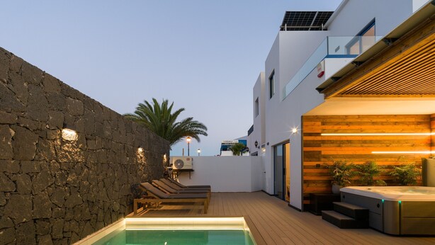 A swimming pool with lounge chairs, a stone wall, wooden deck, and a modern building with a balcony.