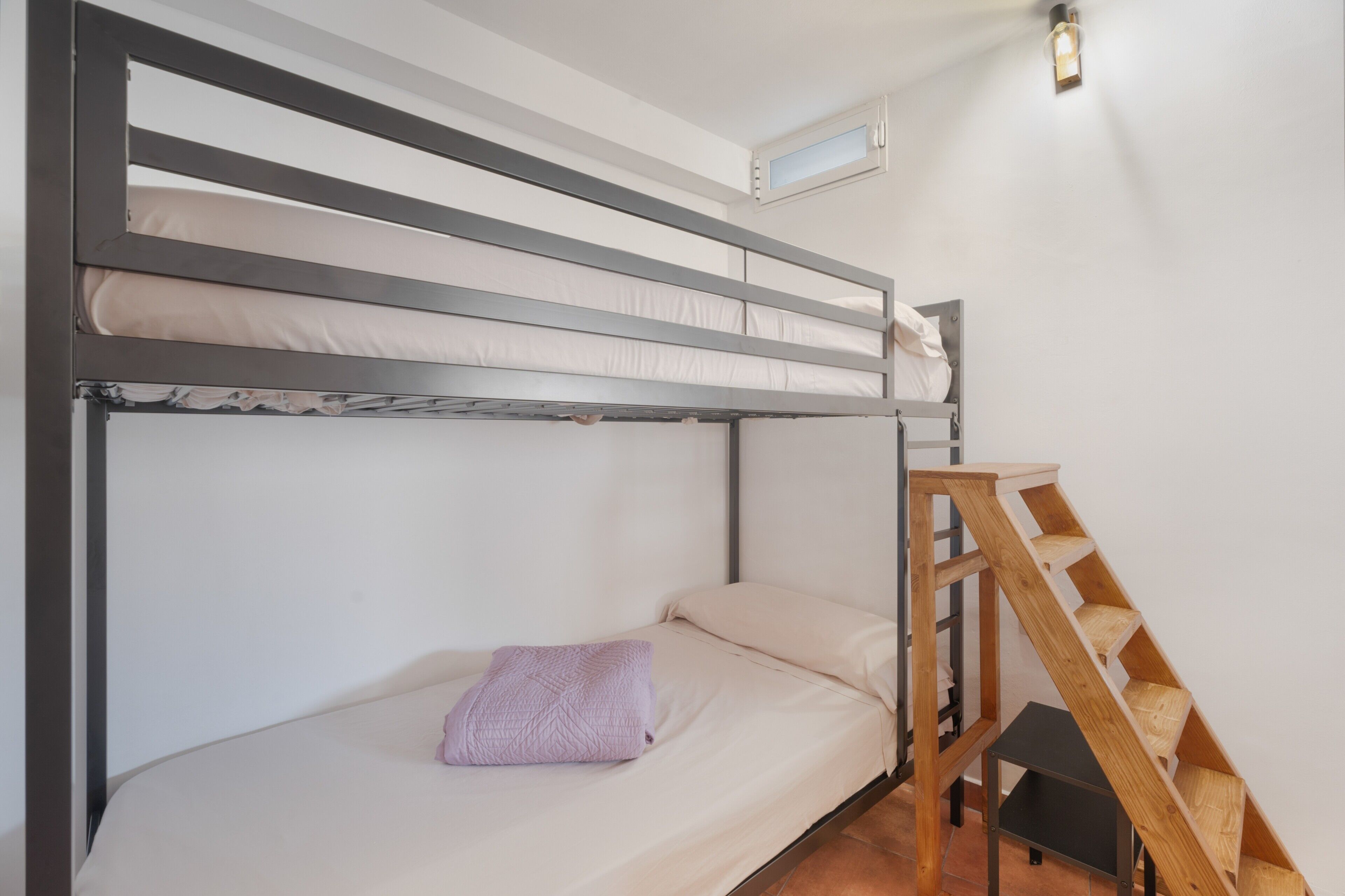A bunk bed with a wooden ladder, a small table, and a window with a light fixture.