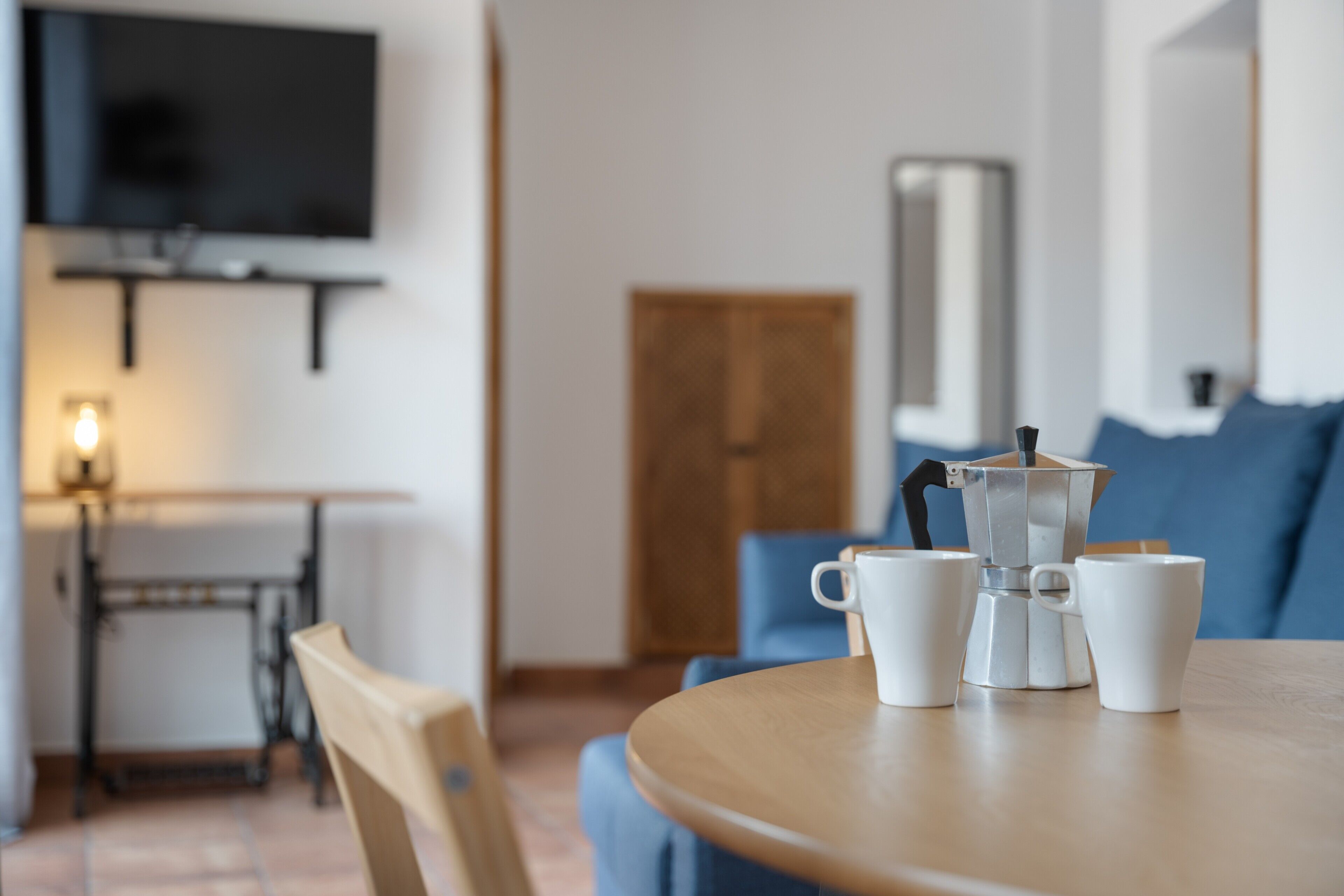Een ronde tafel met een koffiezetapparaat en kopjes, een televisie aan de muur en een deur op de achtergrond.