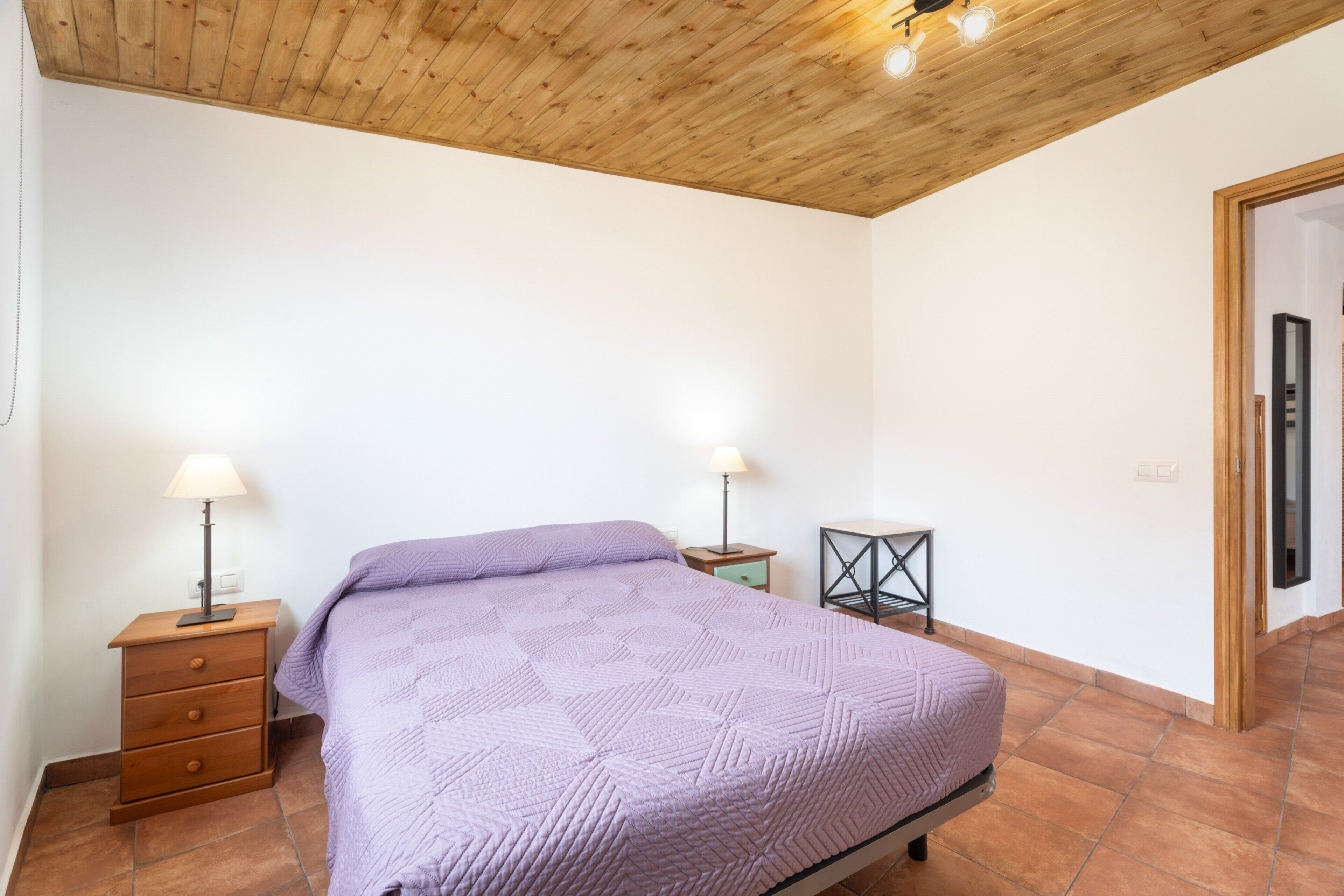 Une chambre à coucher avec un plafond en bois, un lit recouvert d’une courtepointe violette, deux tables de chevet avec des lampes et une petite table également dotée d’une lampe.