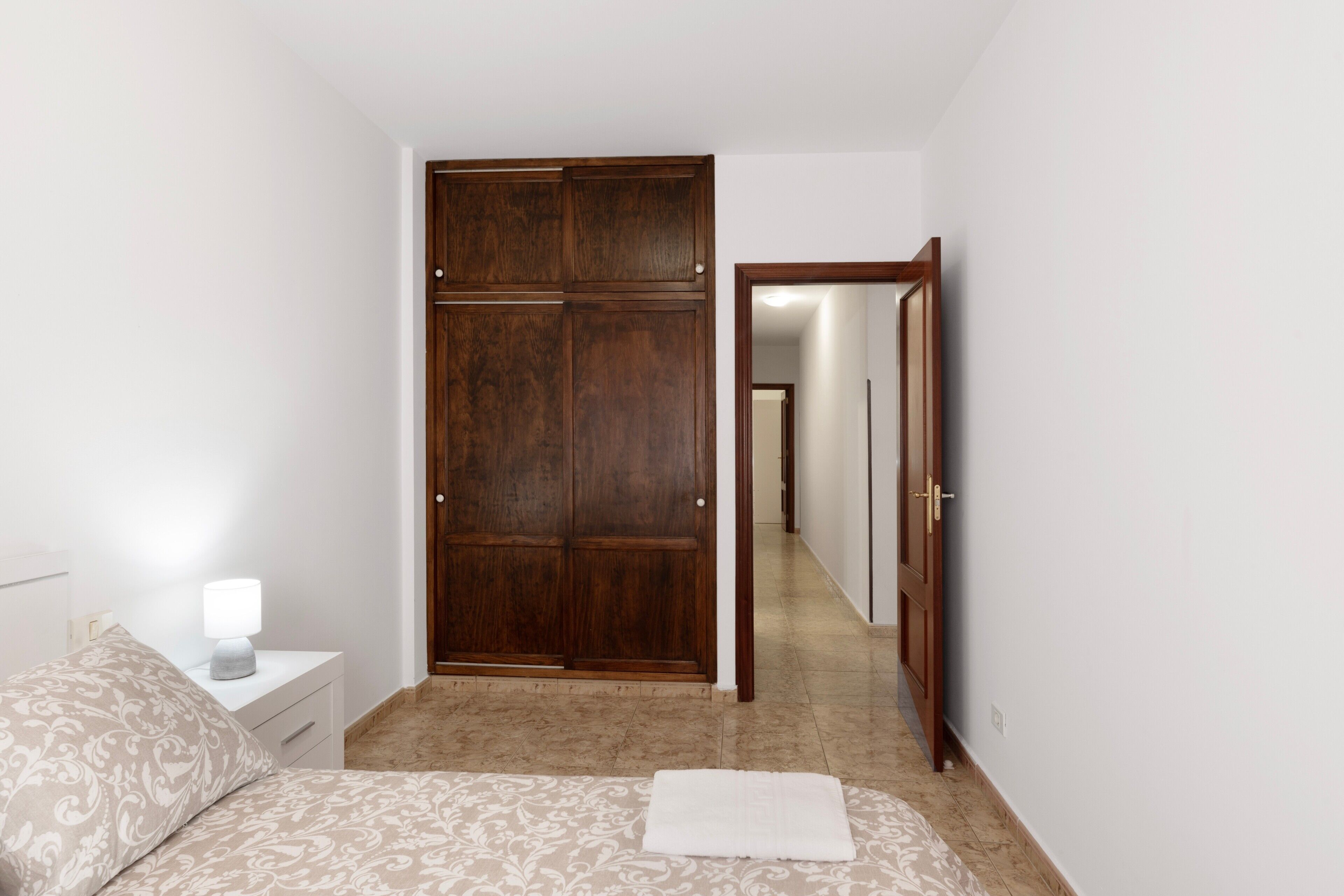 A bedroom with a bed, a nightstand, a lamp, a wooden wardrobe, and a mirror.