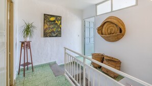 Un pasillo con suelo estampado verde, un atril de madera para plantas, un cuadro y cestas tejidas en una pared blanca.