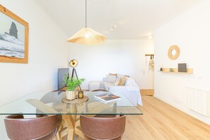 A modern living room with a glass dining table, leather chairs, a sofa, a hanging light fixture, and a framed picture on the wall.