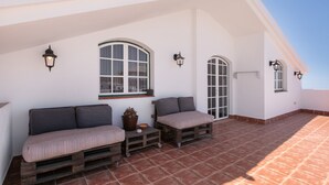 Eine Dachterrasse mit einer weißen Wand, zwei Fenstern mit grüner Verzierung und einer hölzernen Bank mit Kissen.
