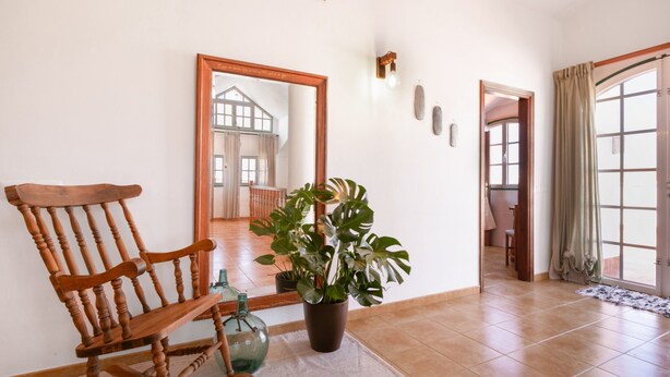 Ein hölzerner Schaukelstuhl, ein Blumentopf, ein Spiegel und ein Fliesenboden in einem Raum mit einem großen Fenster und einer Tür.