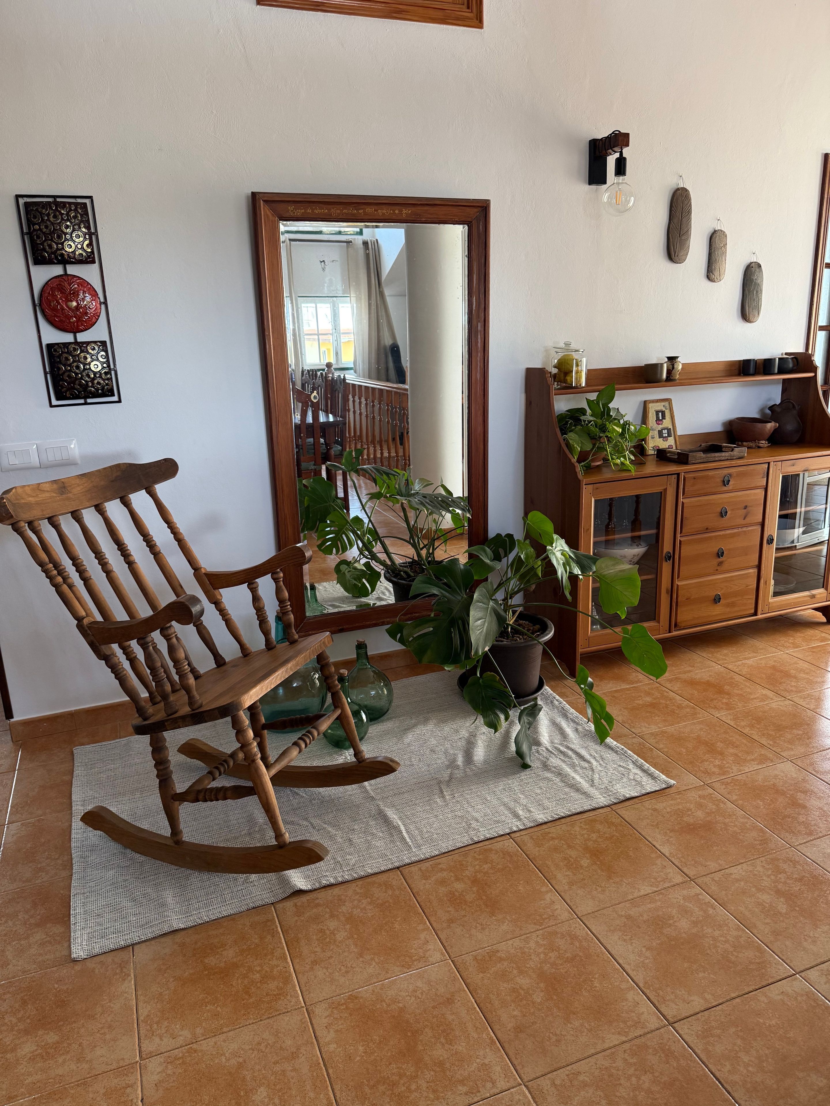 A room with a wooden rocking chair, a large mirror, and a cabinet with plants.