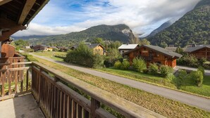 A wooden building with a balcony overlooking a road and houses, with mountains in the background.