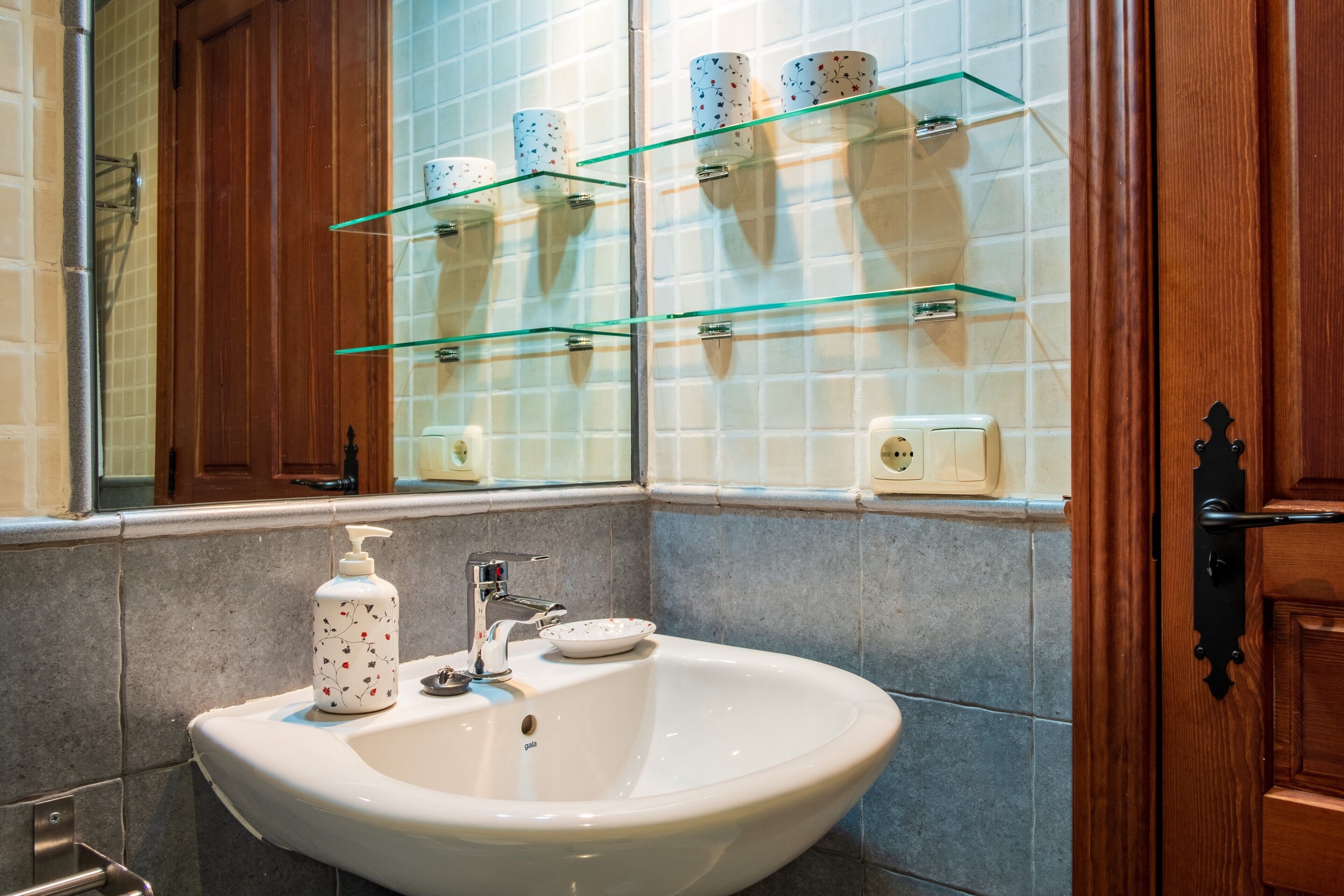 Un baño con un lavamanos blanco, un espejo y estantes de vidrio sobre el lavamanos.
